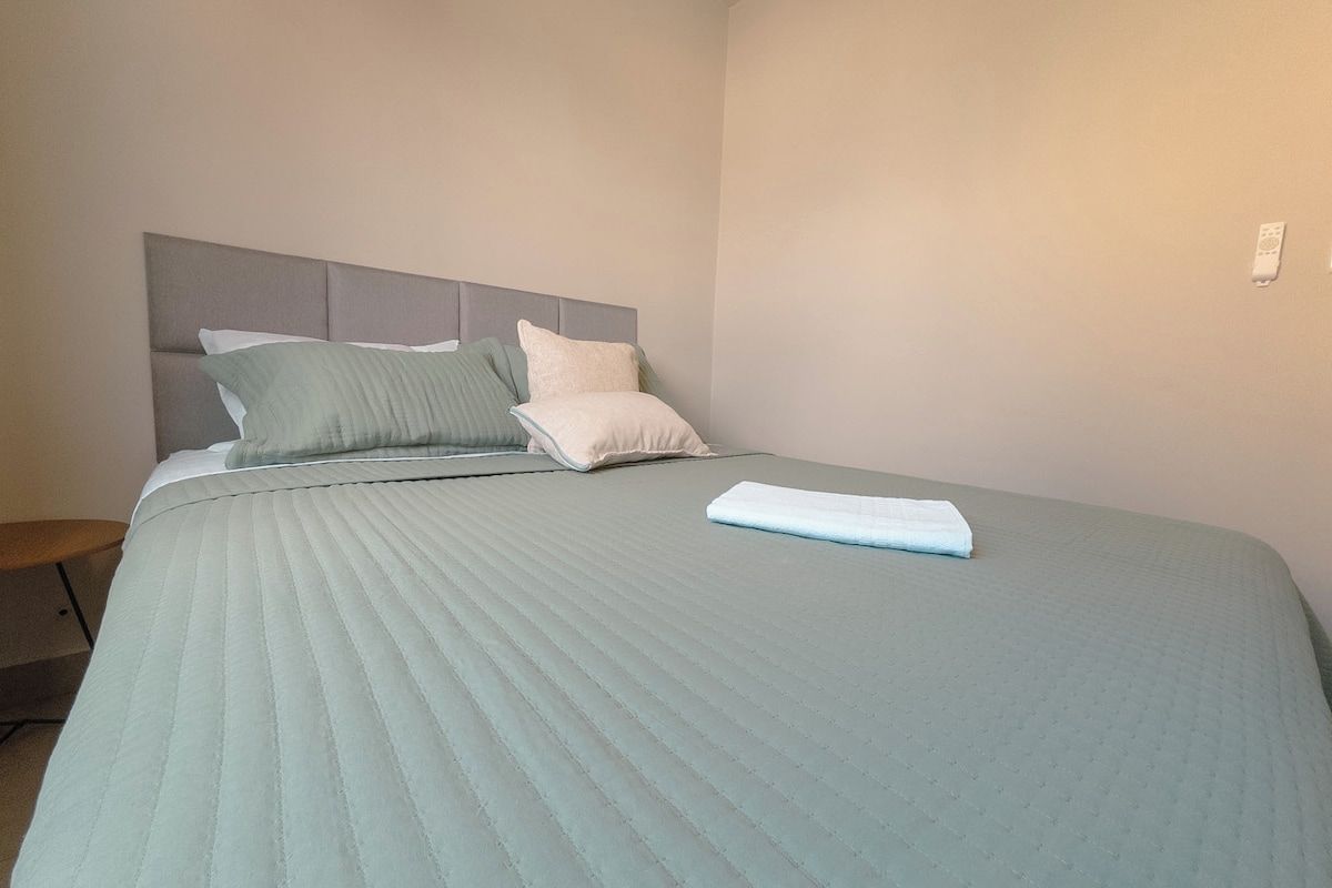 A neatly made bed with a grey headboard and pillows, a folded towel on the bed, and a small wooden stool beside the bed.