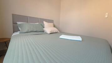 A neatly made bed with a grey headboard and pillows, a folded towel on the bed, and a small wooden stool beside the bed.