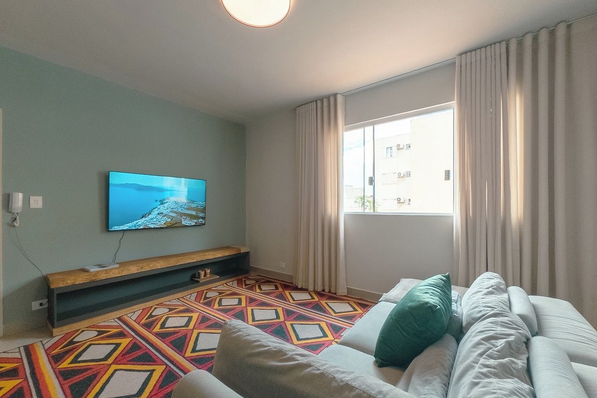 Una sala moderna con una televisión de pantalla plana montada en la pared, una alfombra estampada, un sofá con cojines y una ventana con cortinas.
