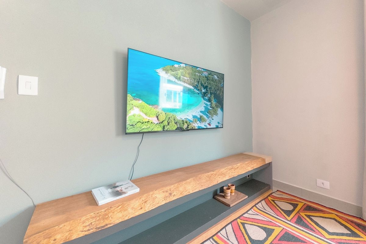 Una habitación con una televisión de pantalla plana montada en la pared, un banco de madera con una revista y una alfombra estampada en el suelo.