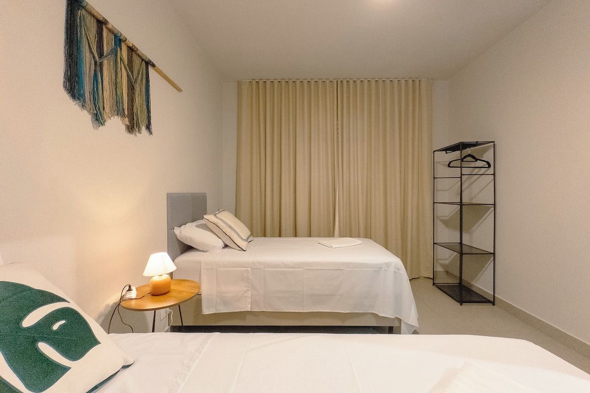 A hotel room with a bed, a bedside table with a lamp, a hanging decoration, a wardrobe, and a curtain.