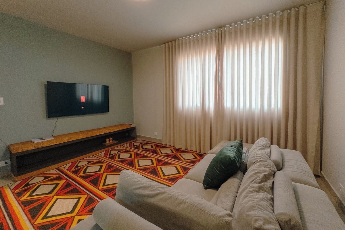 A living room with a sofa, a flat-screen TV mounted on the wall, a patterned rug, and a window with curtains.