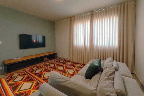 A living room with a sofa, a flat-screen TV mounted on the wall, a patterned rug, and a window with curtains.