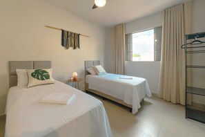 Una camera d'albergo con due letti, un tavolino con una lampada, una finestra con tende e un appendiabiti.