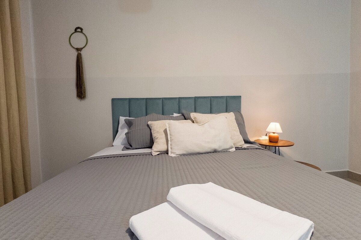 Cama bem arrumada com colcha cinzenta e almofadas, mesa de cabeceira com abajur e porta-chaves fixado na parede.