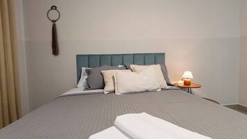 Cama bem arrumada com colcha cinzenta e almofadas, mesa de cabeceira com abajur e porta-chaves fixado na parede.