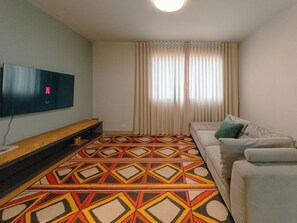 A living room with a flat-screen TV, a sofa, a patterned rug, and a window with curtains.
