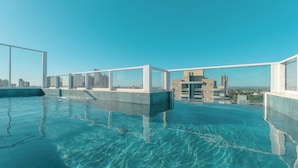 Ein Pool ohne erkennaren Rand auf dem Dach mit klarem, blauem Wasser, umgeben von Glasgeländern und modernen Gebäuden im Hintergrund.