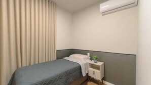 Ein Einzelbett mit grauer Decke, ein Nachttisch mit Pflanze und eine wandmontierte Klimaanlage.