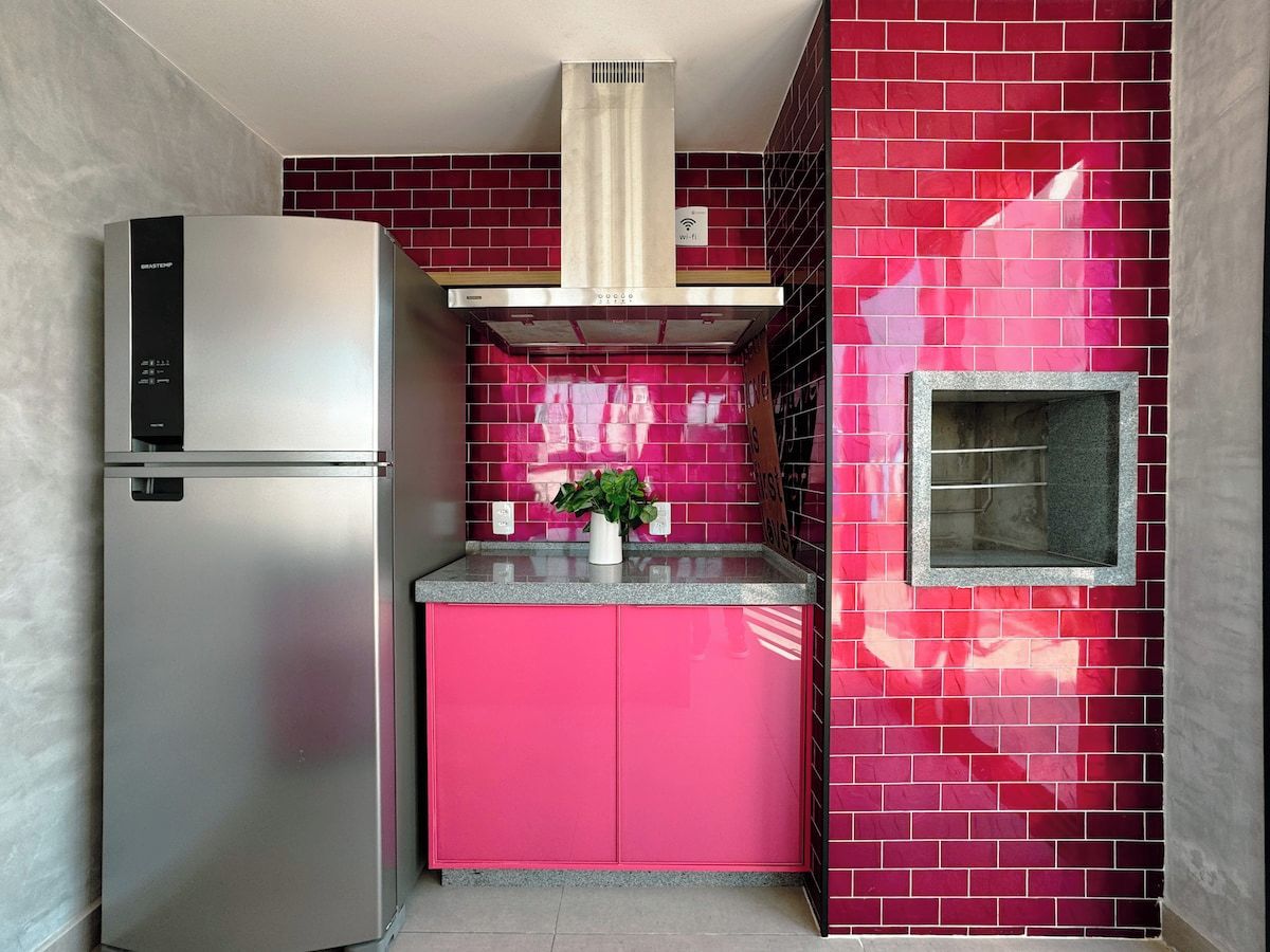 A compact kitchen with a stainless steel refrigerator, a pink cabinet, and a red-tiled backsplash.