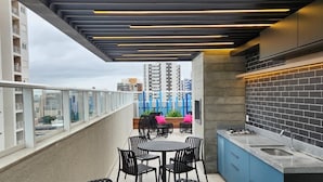 Eine Dachterrasse mit einem runden Tisch und Stühlen, einer Küchenzeile mit Spüle sowie Blick auf Gebäude im Hintergrund.