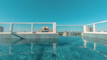 A rooftop infinity pool with clear blue water, surrounded by a white railing, and a view of distant buildings.