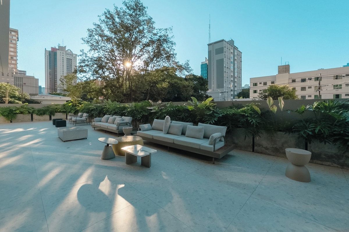 Um lounge no terraço com mobiliário externo, uma grande árvore e edifícios da cidade ao fundo.