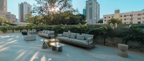 A rooftop lounge with outdoor furniture, a large tree, and city buildings in the background.