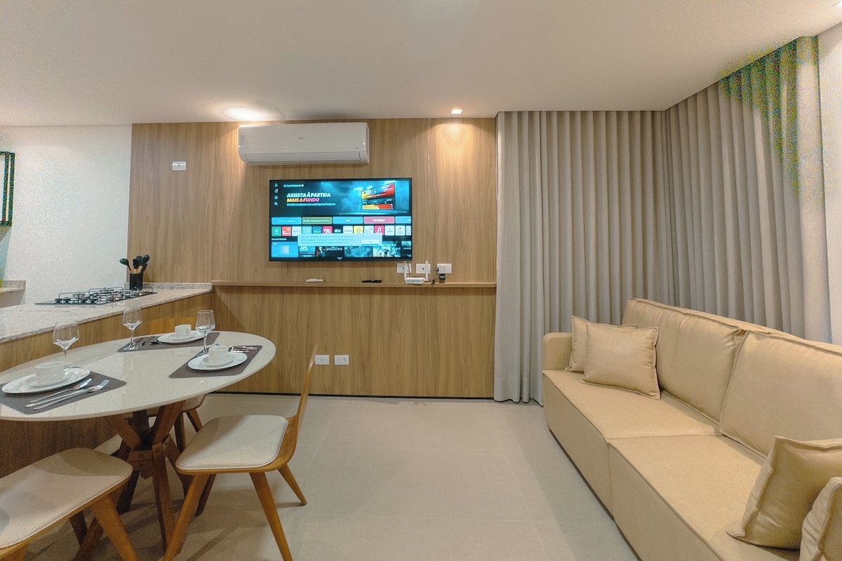 A modern living room with a flat-screen TV mounted on the wall, a beige sofa, and a dining area with a round table and chairs.