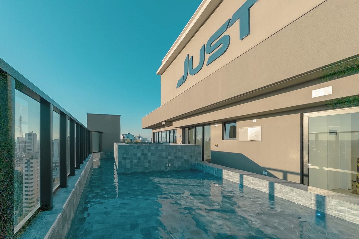 Piscina no terraço com barreira de vidro, um edifício com o letreiro 'JUST' e vista para a cidade.