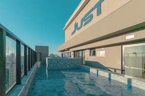 A rooftop swimming pool with a glass barrier, a building with a 'JUST' sign, and a cityscape view.