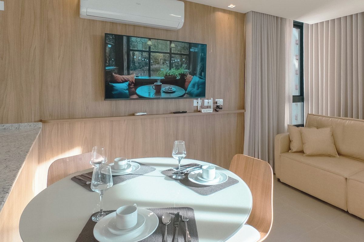 A modern living room with a dining area, a flat-screen TV mounted on the wall, and a sofa.