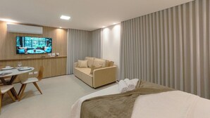Quarto de hotel com cama, sofá, televisão e área de refeições.
