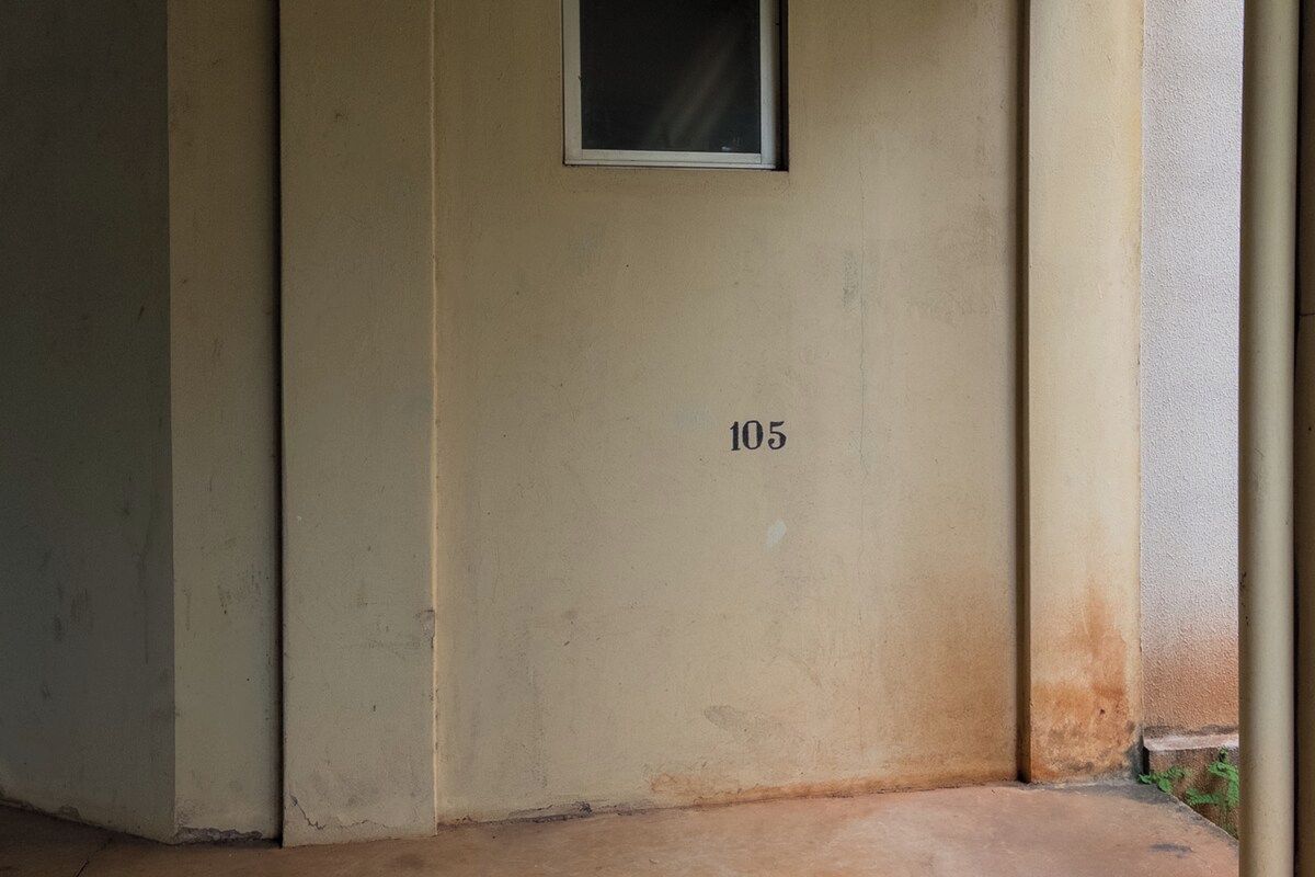 Un mur de couleur beige portant le numéro 105, une petite fenêtre et un sol en béton.