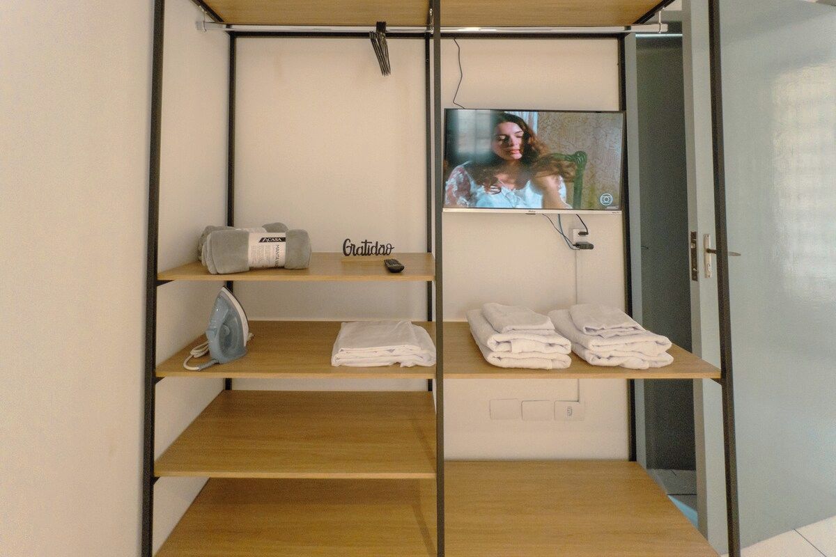 Habitación de hotel con televisor en la pared, estante con toallas dobladas y un plancha portátil.