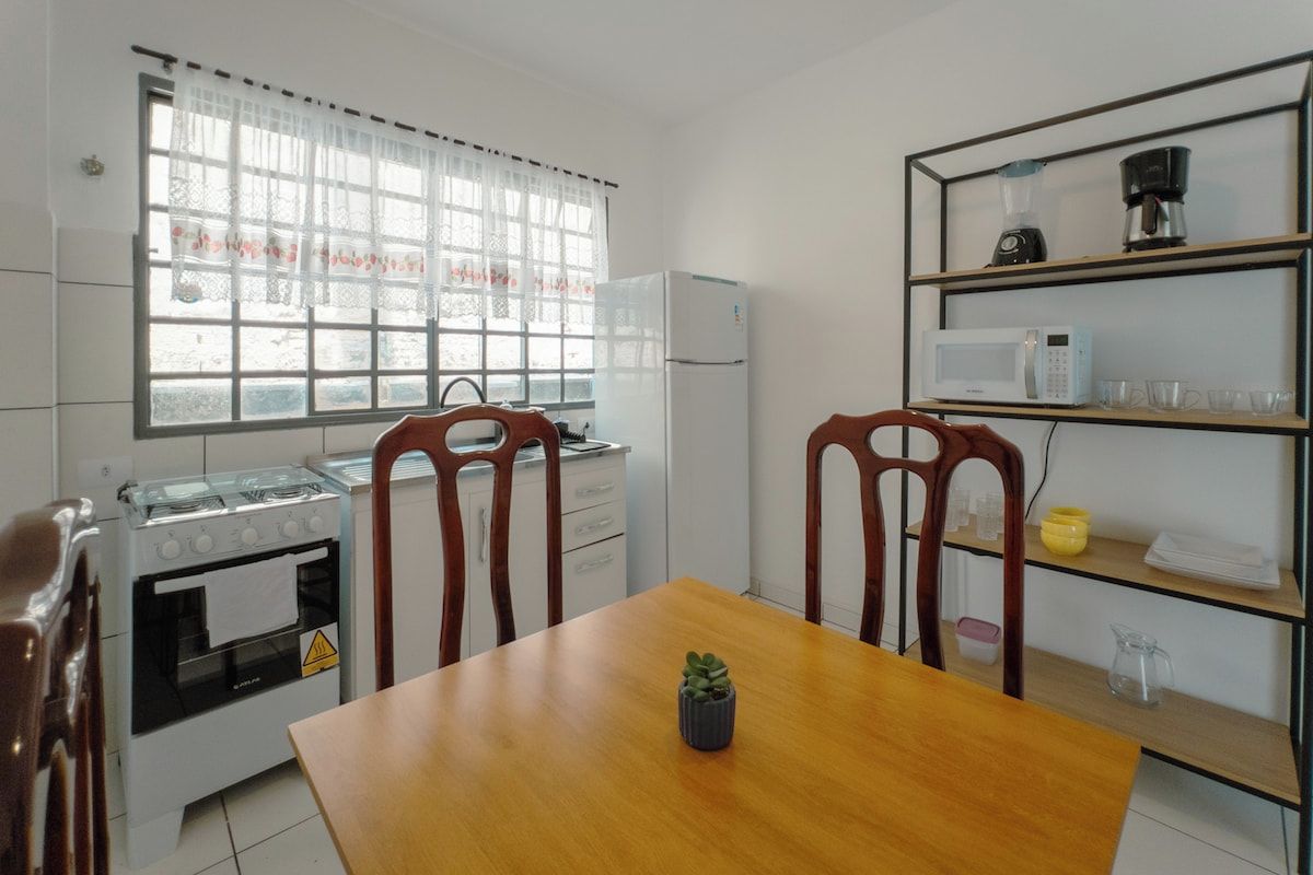Cocina compacta con estufa, microondas y refrigerador. Hay una mesa de comedor con sillas y una pequeña planta en maceta.