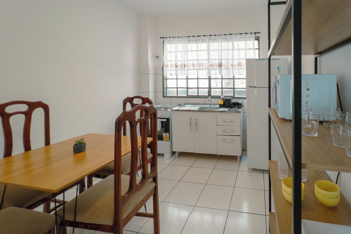 Cocina compacta con armarios blancos, una ventana con cortinas y una zona de comedor con mesa y sillas de madera.