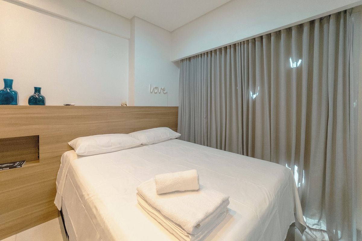Een hotelkamer met een net opgemaakt bed, een houten hoofdbord, een wandplank met decoratieve objecten, en een groot raam met gordijnen.