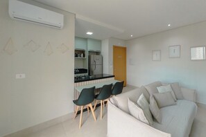 Una sala moderna con un sofá y sillones, una cocina con refrigerador y estufa, y una mesa de comedor con sillas.