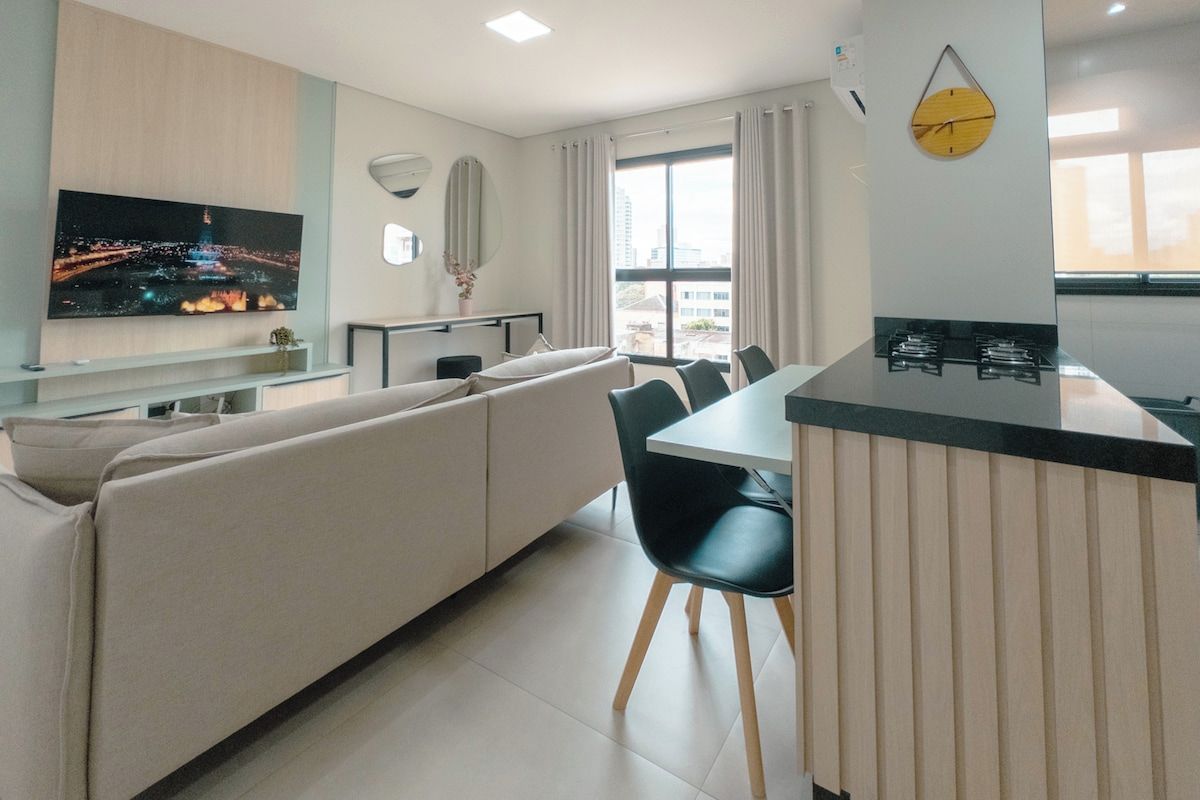 Una sala moderna con sofá, una mesa de comedor con sillas, un área de cocina con estufa y un ventanal con cortinas.