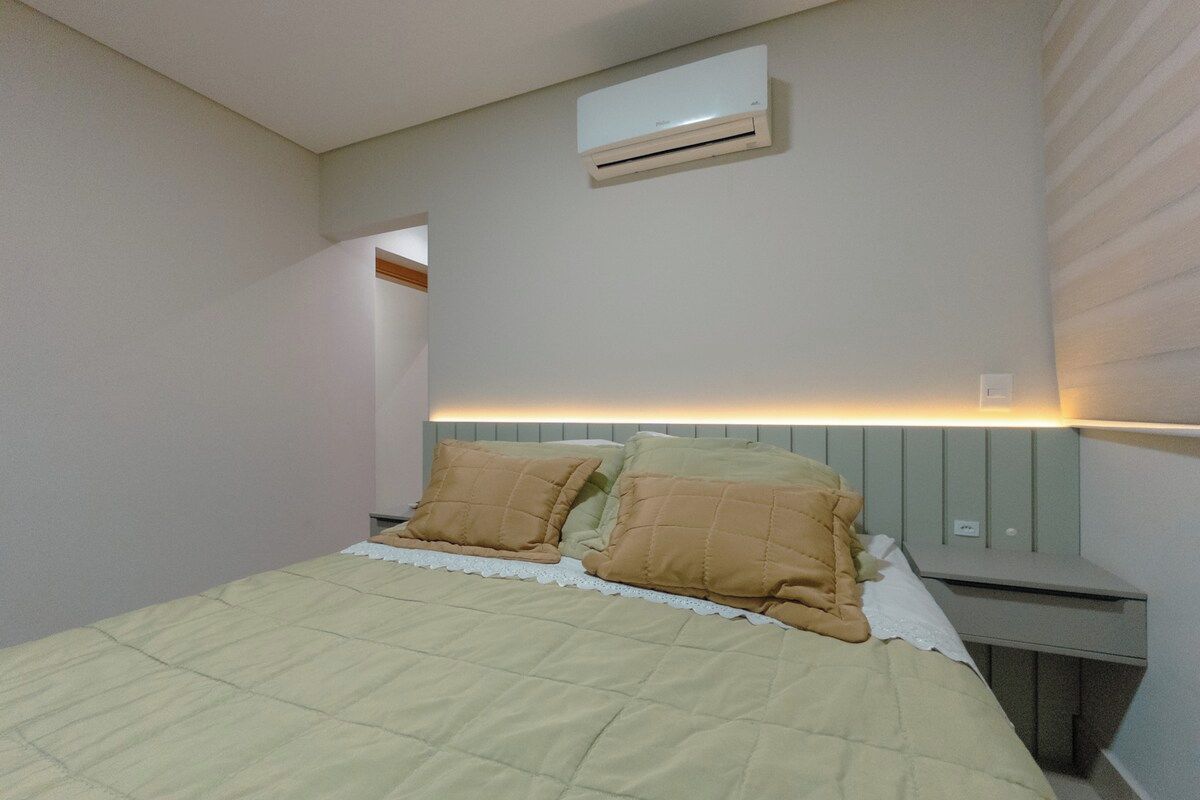 A bedroom with a bed, two pillows, a nightstand, an air conditioning unit, and a wall-mounted light.