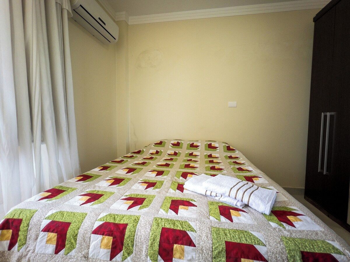 Un lit recouvert d’une courtepointe à motifs, une serviette blanche, une armoire sombre et une fenêtre avec des rideaux.