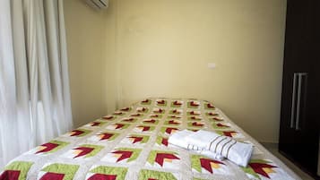 Un lit recouvert d’une courtepointe à motifs, une serviette blanche, une armoire sombre et une fenêtre avec des rideaux.