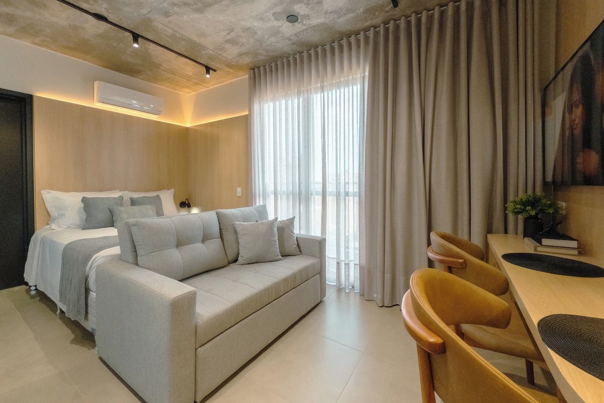 A modern hotel room with a bed, sofa, dining table, chairs, and a large window with curtains.