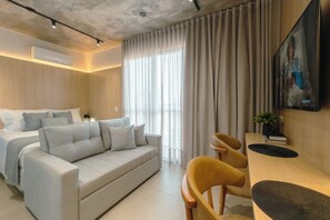 Quarto de hotel moderno com cama, sofá, mesa de jantar e uma grande janela com cortinas.
