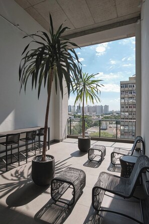 Uma varanda com palmeira em vaso, mesa de jantar com cadeiras e vista para a paisagem urbana.