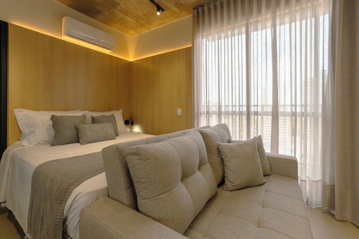 A hotel room with a bed, a sofa, a window with sheer curtains, and an air conditioning unit.