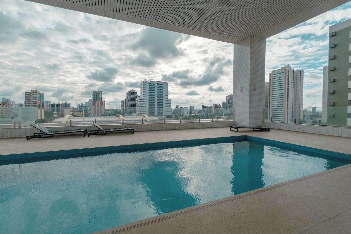 Ein Pool auf dem Dach mit Blick auf die Stadtlandschaft, umgeben von einer gefliesten Terrasse und Liegestühlen.