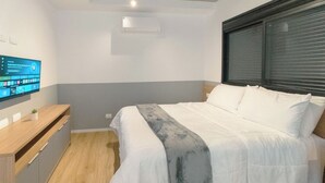 Quarto com cama, televisão fixada na parede, cómoda de madeira e janela com persianas.