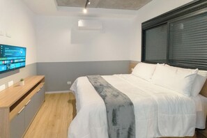 Een slaapkamer met een bed, een aan de muur bevestigde televisie, een houten dressoir en een raam met jaloezieën.