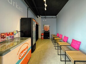 Ein Café mit einer Theke, auf der ein Geben-Produkt präsentiert wird, einer Sitzgelegenheit mit Tischen und rosafarbenen Kissen sowie einem Kühlschrank im Hintergrund.