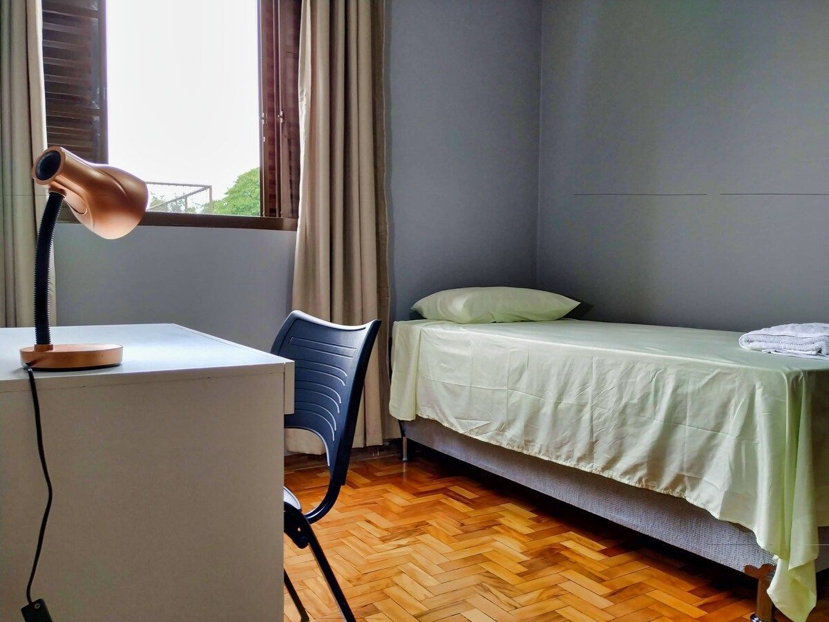 A room with a bed, a desk with a lamp, a chair, and a window with curtains.