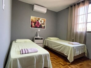 A room with two beds, a nightstand, an air conditioner, a painting, and a window with curtains.