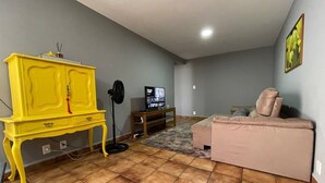 Uma sala de estar com um armário amarelo, uma televisão, um sofá rosa e um chão em mosaico.