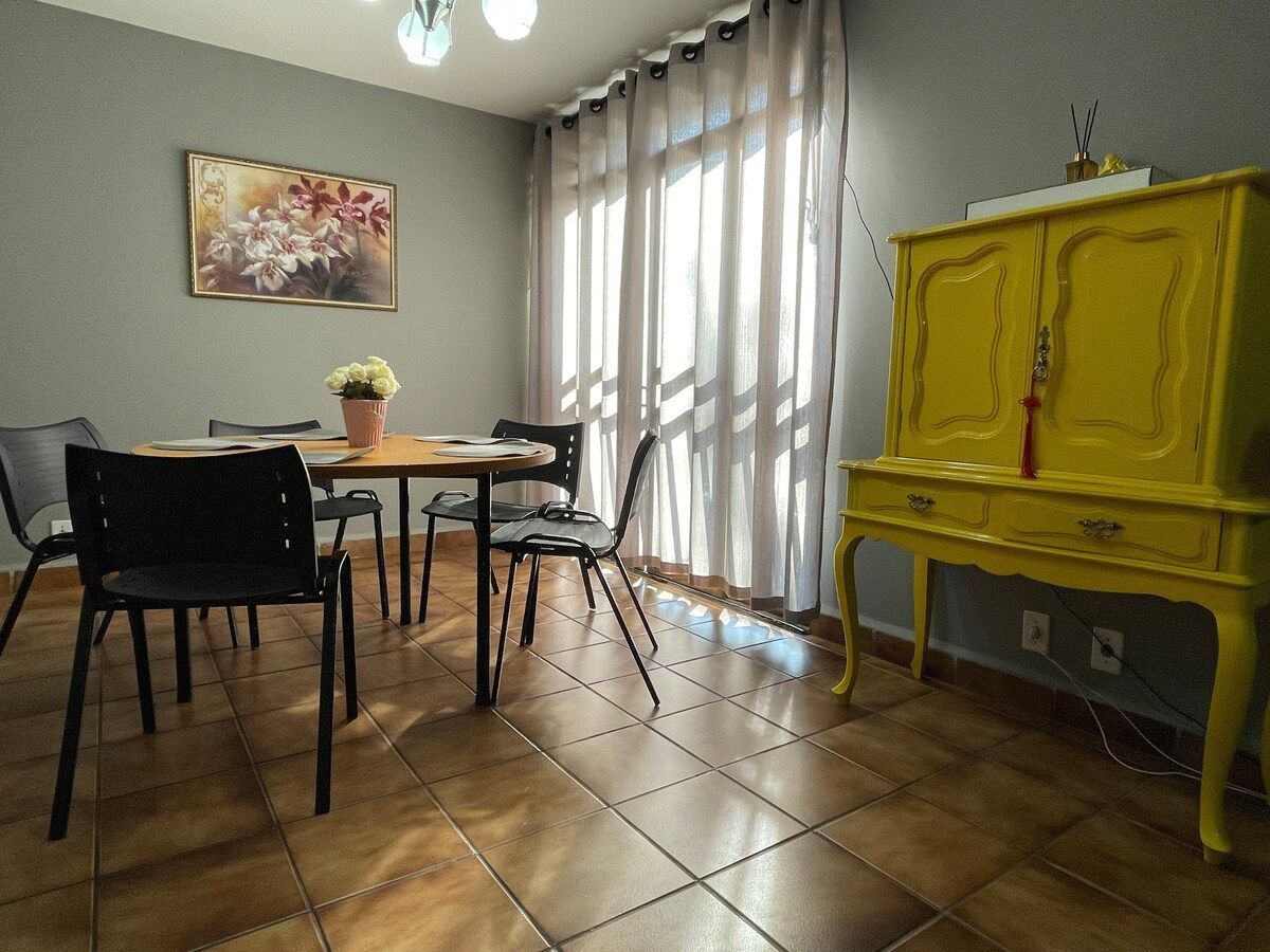 Een eetgedeelte met een tafel, stoelen, een geel kastje en een schilderij aan de muur.
