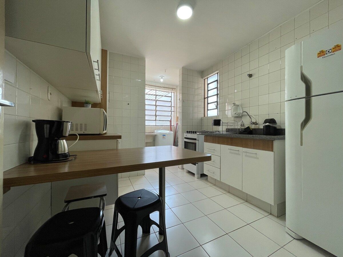 Una cocina con armarios blancos, encimera de madera, cafetera, refrigerador y ventana con persianas.
