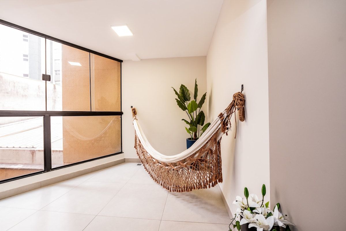 A room with a large window, a hammock, a potted plant, and a wall-mounted decoration.