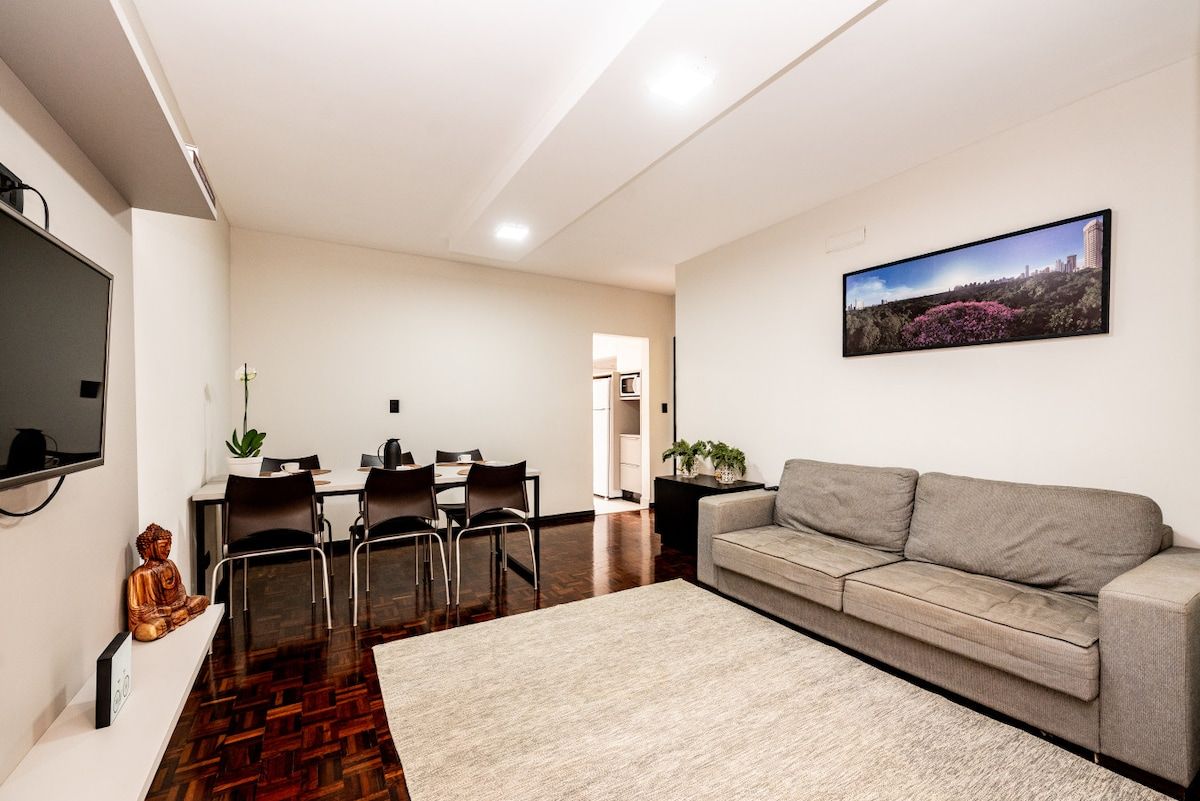 A modern living room with a sofa, a dining area with chairs and a table, a flat-screen TV mounted on the wall, and a framed picture above the sofa.