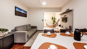 Ein modernes Wohnzimmer mit einem grauen Sofa, einem Esstisch mit zwei Stühlen, einer Hängematte und einem an der Wand befestigten Fernseher.