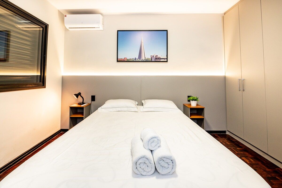A hotel room with a large bed, two bedside tables, a wall-mounted air conditioner, a framed picture, and a wardrobe.
